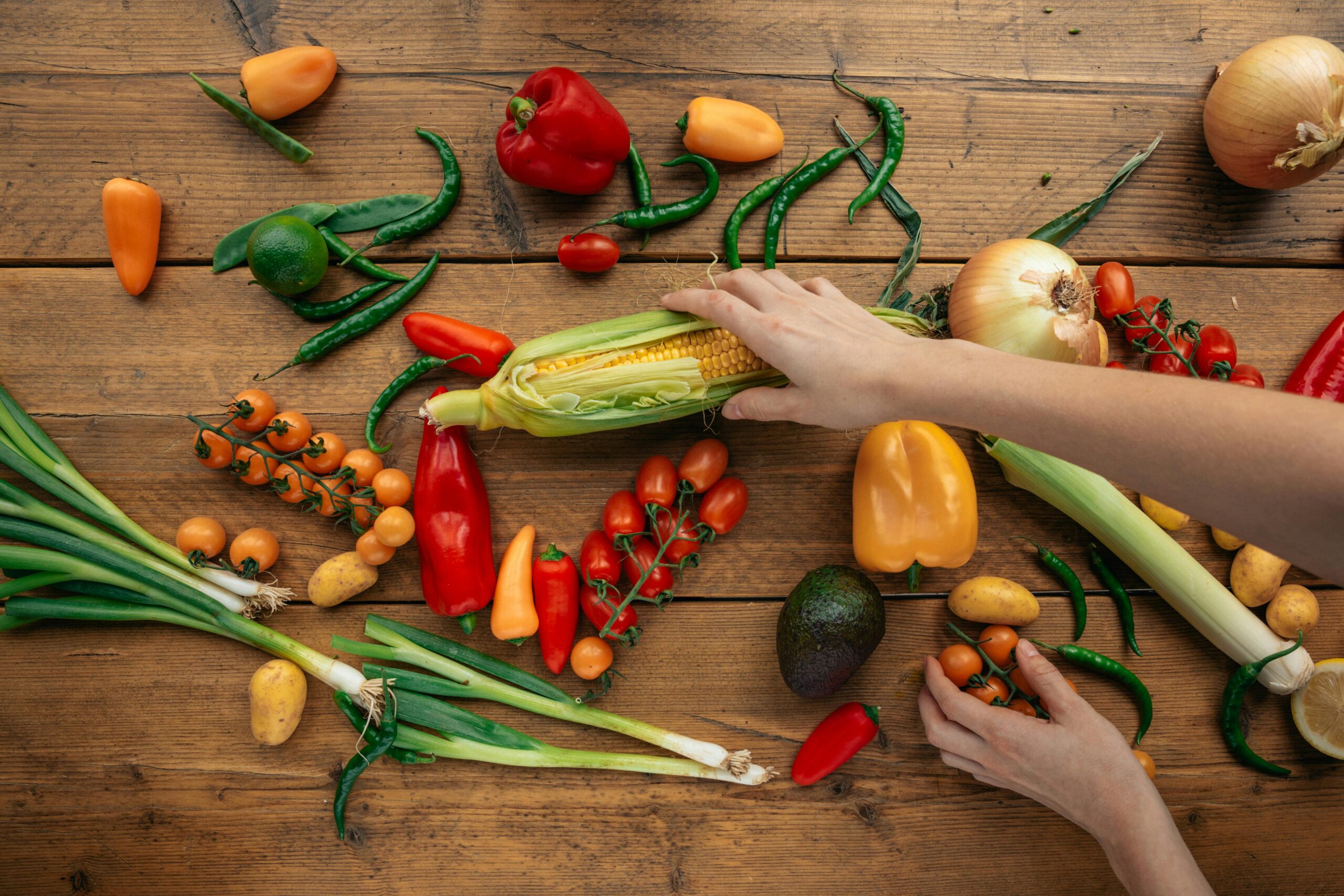 Bright assortment of fresh vegetables on a wooden table, featuring hands arranging them.